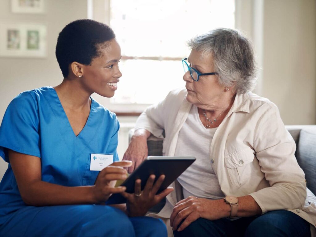 A nurse and an older woman sit together on a couch, engaged in a conversation, conveying a sense of care and companionship.